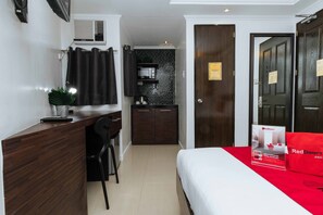 A hotel room with a wooden desk, a small kitchenette, a bed with a red pillow, and a dark wooden door. - RedDoorz Plus @ Poblacion Makati (Makati)