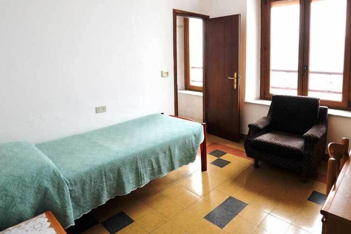A small, simple room with a single bed, a chair, and a door leading to another room.