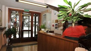 A reception area with a wooden reception desk, a potted plant, and a sign indicating the entrance to the building. - Hotel Conca D'Oro (Lama Mocogno)