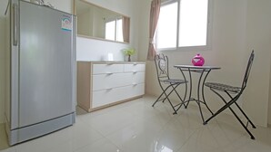 A compact kitchen with a refrigerator, a white cabinet, a dining table with chairs, and a window with curtains. - Sansuk Apartment (Pathum Thani)