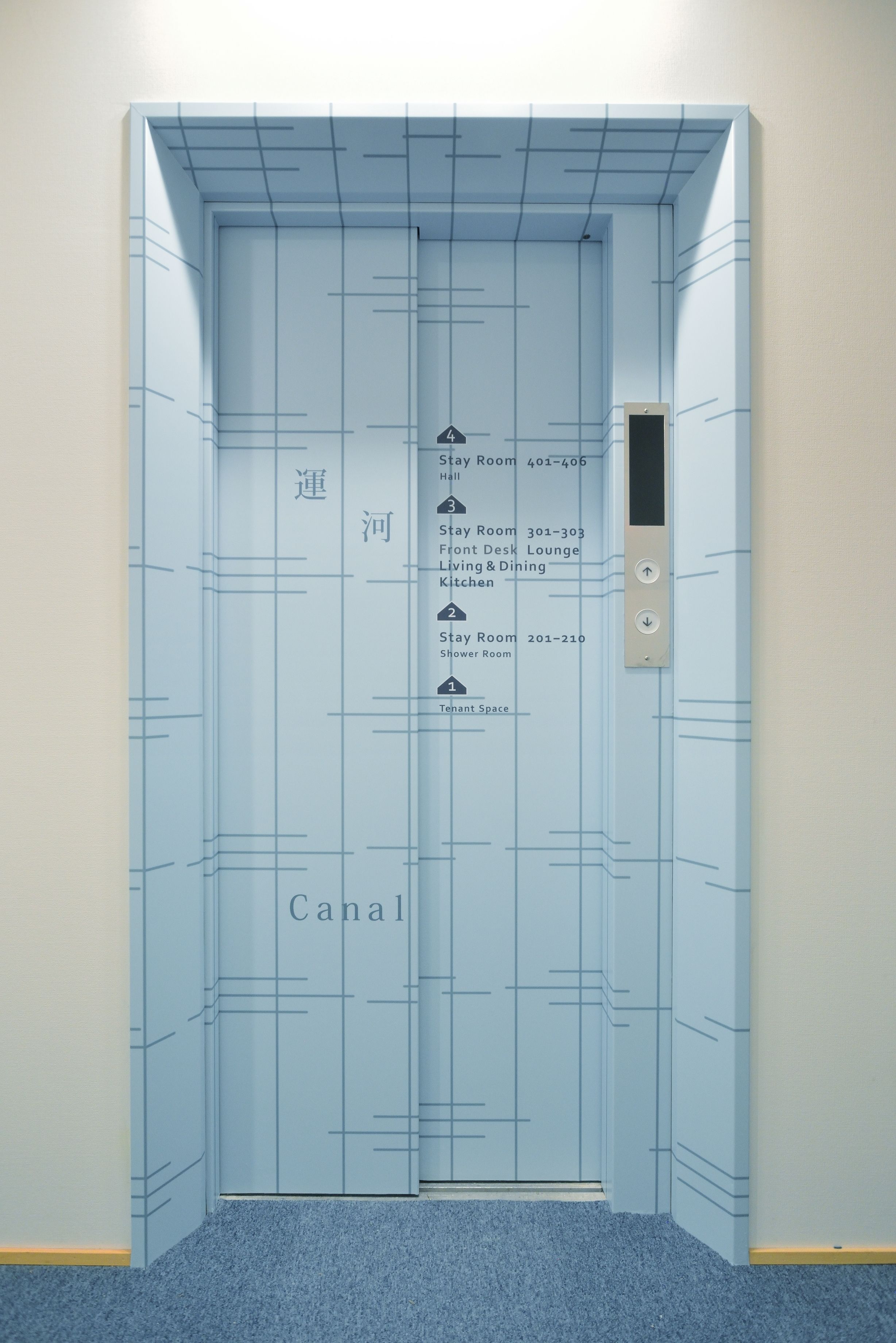 Un ascensor con un estampado azul a cuadrícula y una pantalla digital. Tiene un cartel que muestra números de habitaciones y nombres como Habitación Stay 401-406, Salón de Recepción y Cocina.