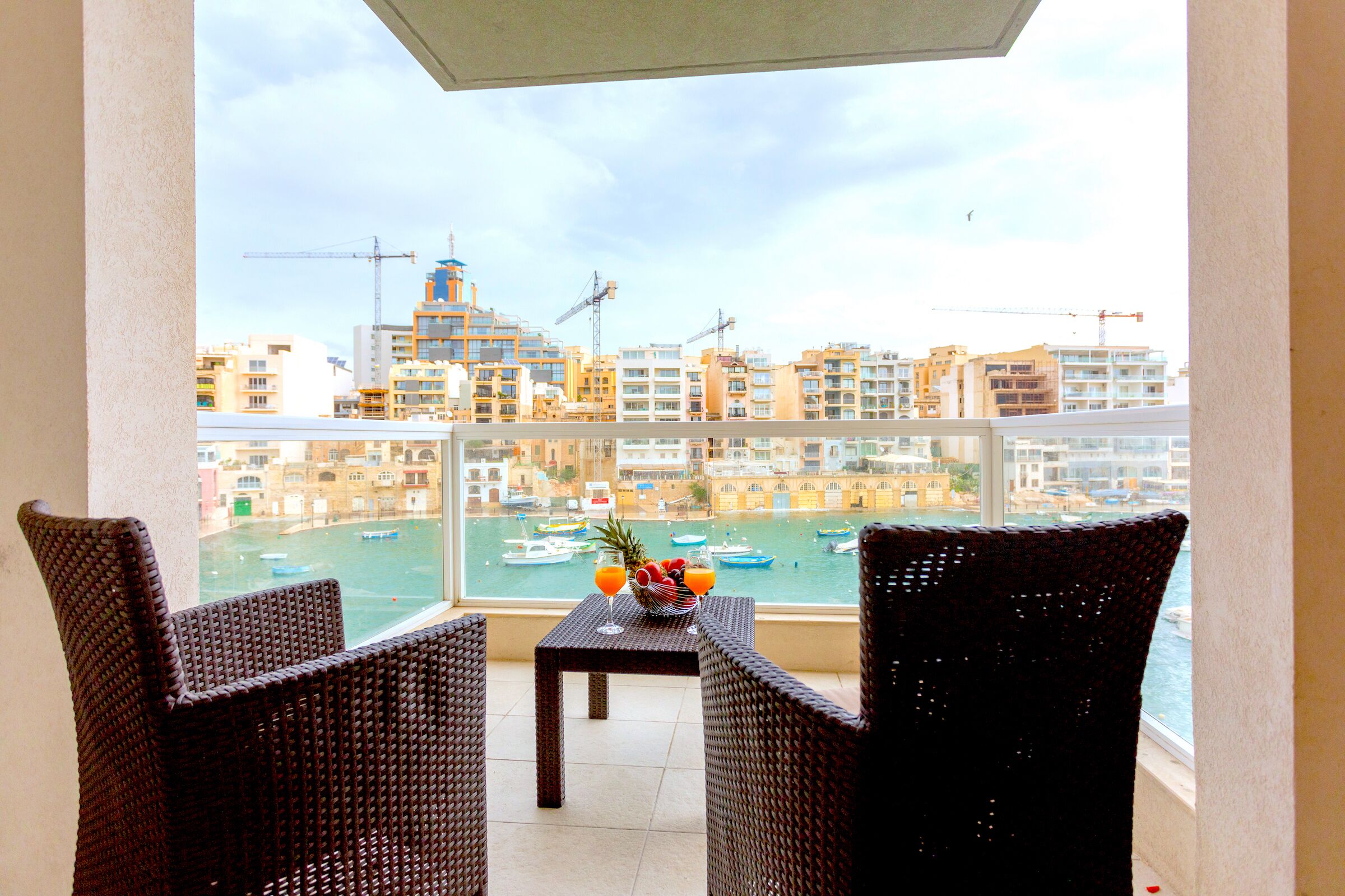 Un balcón con sillas de mimbre y una mesa con fruta y un vaso. Se ve una marina con barcos y edificios a través de la ventana.