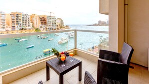 A balcony with a view of the sea, a table with fruit and drinks, and wicker furniture.