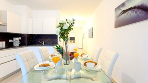 A modern dining area with a glass table, white chairs, and breakfast items including croissants, orange juice, and a fruit bowl.