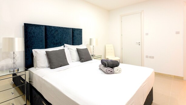 A neatly made bed with white and gray linens, a dark blue headboard, and a bedside table with a lamp.