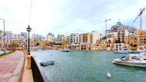 Un paseo marítimo con barcos amarrados, una acera peatonal y edificios al fondo.