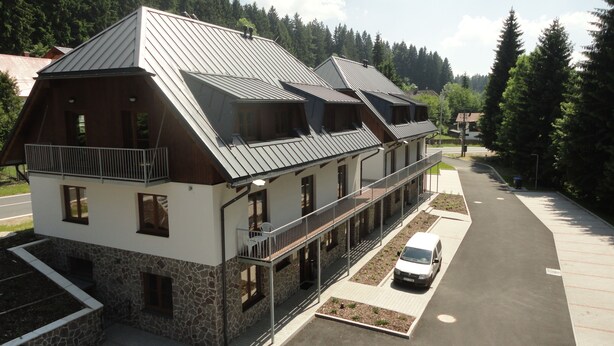 A two-story building with a stone facade and a metal roof, featuring balconies and a parking area.
