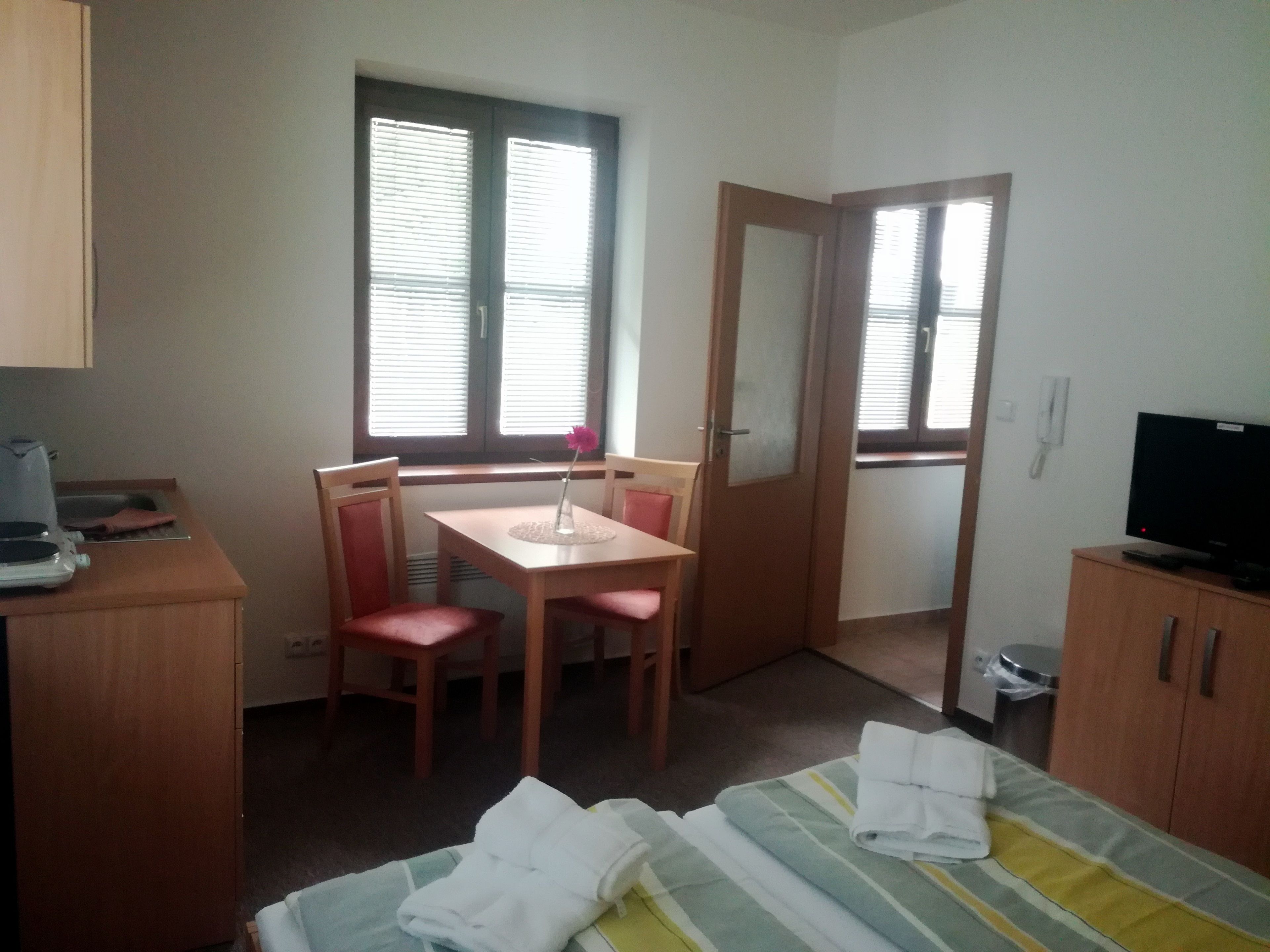 A small, tidy room with a bed, a desk, two chairs, a television, and a window with blinds.