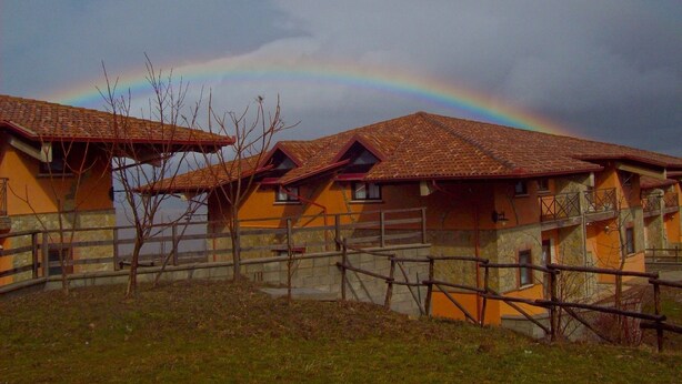 A row of yellow buildings with red-tiled roofs, a wooden fence, and a rainbow in the sky. - Hotel Pietrapanna (Calvello)