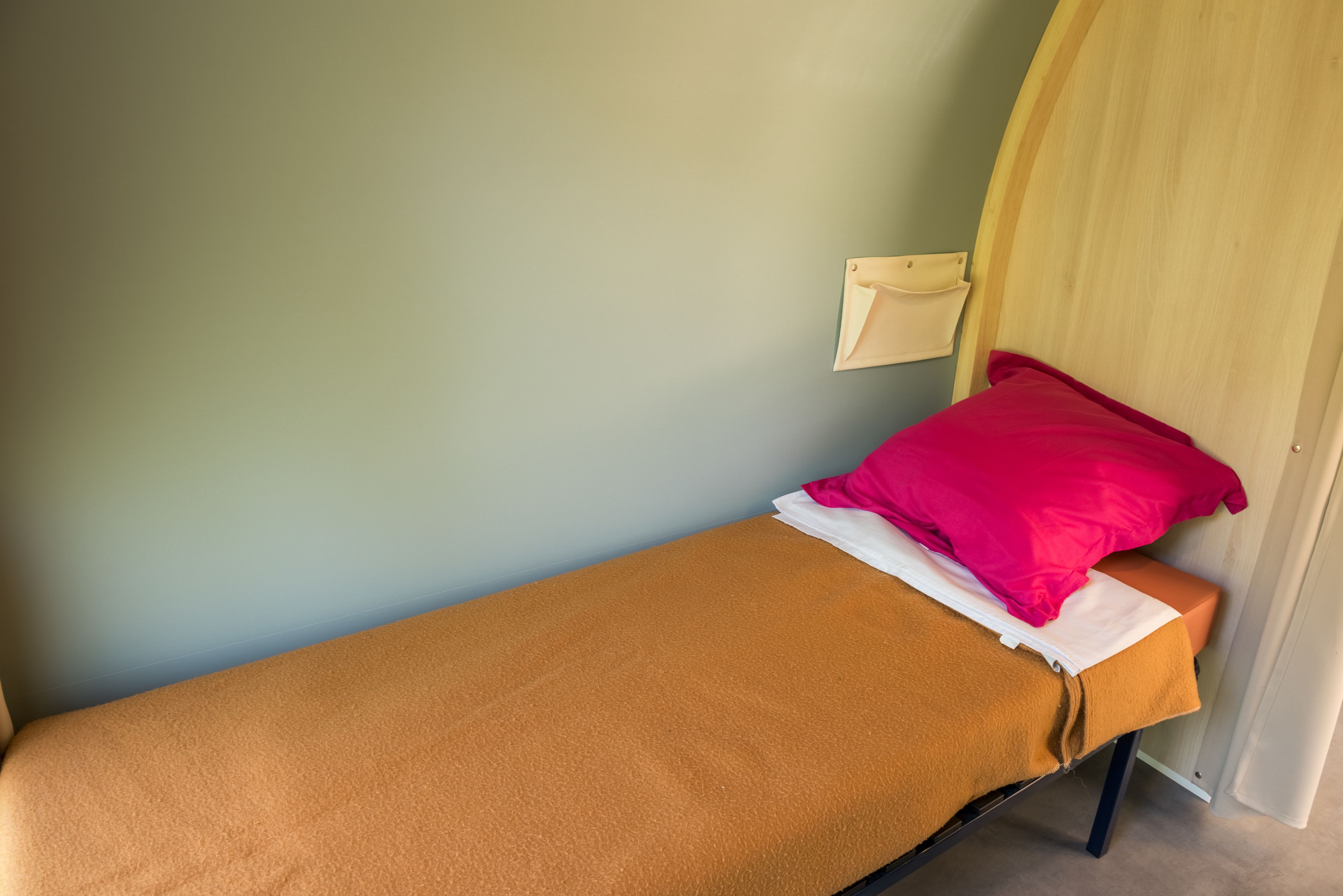 A single bed with a brown mattress and a red pillow in a room with light green walls and a wooden headboard.