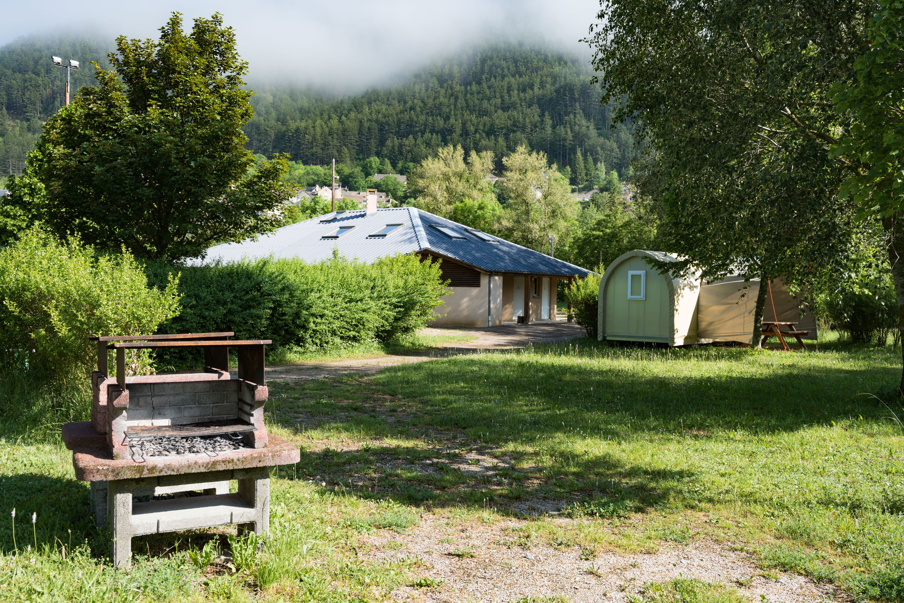 Un cadre extérieur rustique comprenant un barbecue en pierre, un petit abri de jardin et une maison avec un toit en pente, le tout situé dans une zone boisée.
