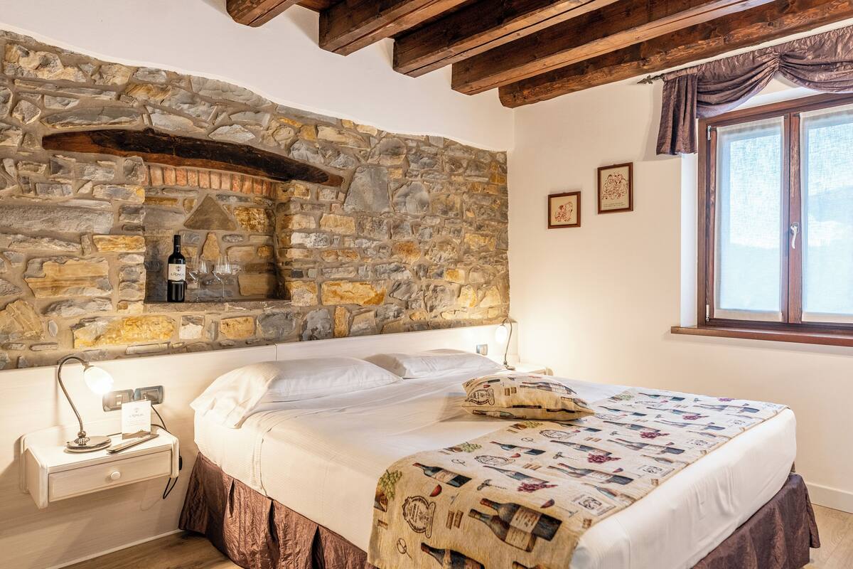 A bedroom with a stone wall, a bed with a patterned blanket, a nightstand with a lamp, and a window with curtains.