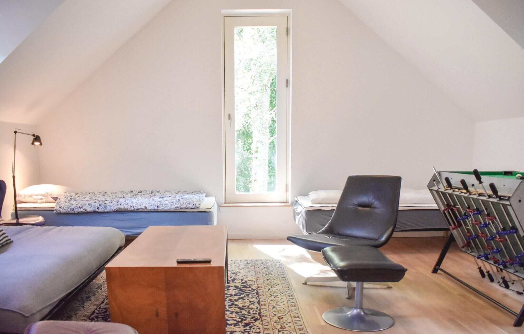Een knus slaapkamerloft met een schuine kap, een groot raam, een bed met beddengoed met een patroon, een zwarte stoel, een kleine tafel en een futon.
