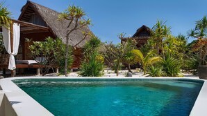 Una piscina cristalina con borde curvo, rodeada de exuberante vegetación tropical y edificios con techo de paja.