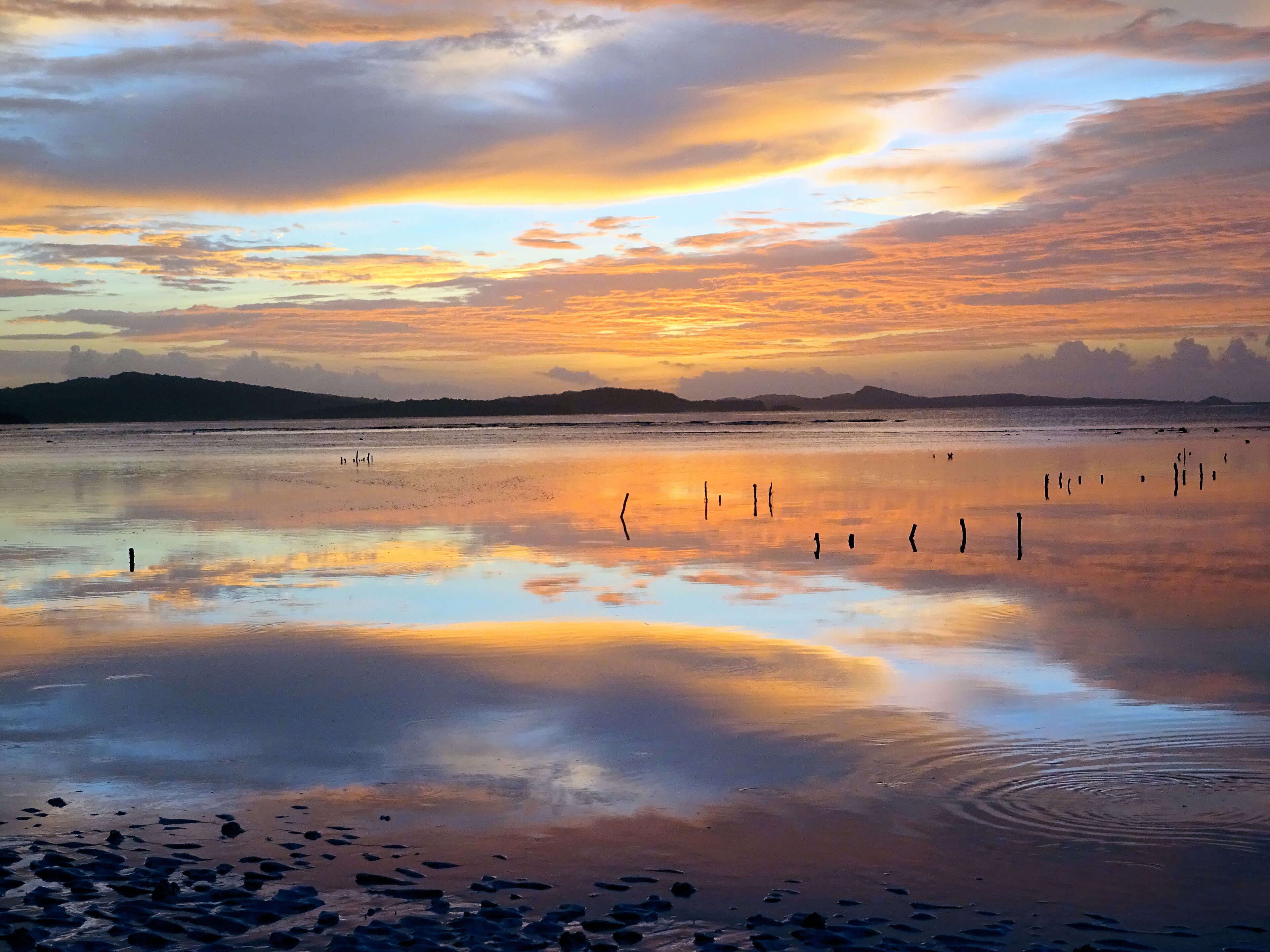 Una playa con aguas tranquilas que reflejan el cielo, una cadena montañosa distante y algunos objetos dispersos en la orilla.
