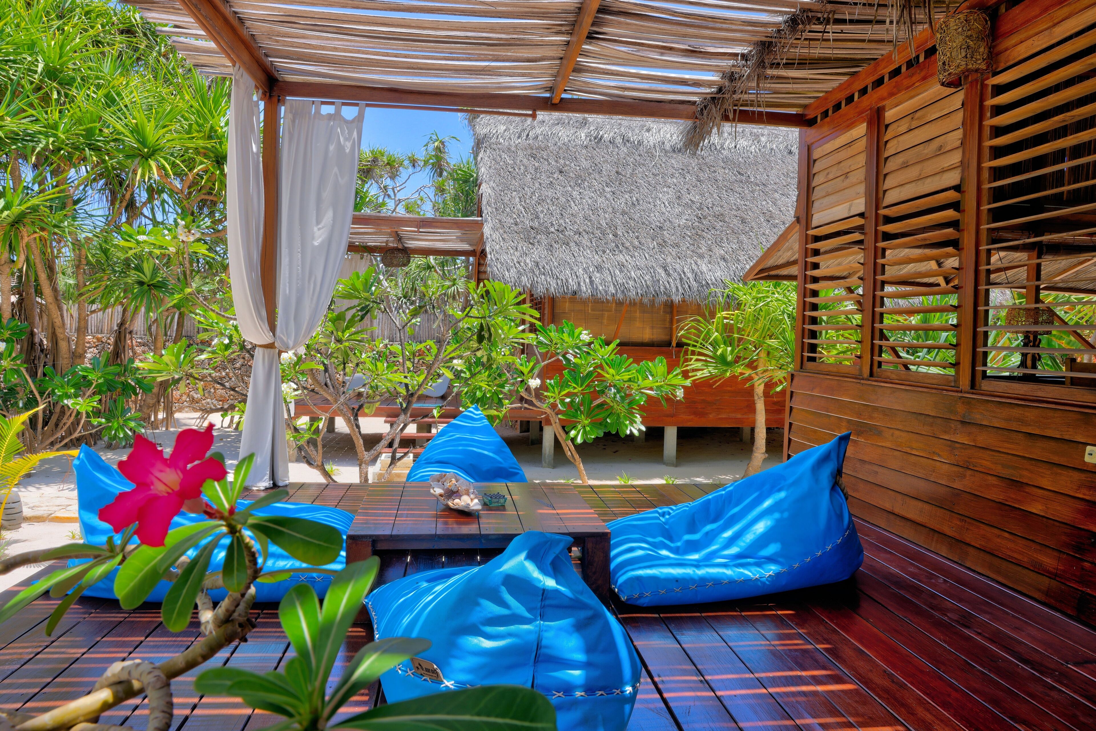 A wooden deck with blue bean bag chairs, a table with a decorative item, and a thatched roof structure.