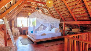 Una habitación de madera acogedora con una cama bajo techo de paja, un sillón colgante y una cesta tejida.