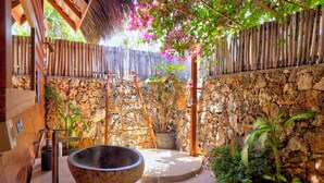 Un baño con paredes de piedra, un recipiente grande, una escalera de madera y un techo de paja.