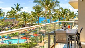 Un balcón con barandilla de vidrio que da a una zona de piscina, palmeras y vistas al océano.