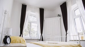 A bedroom with a bed, a nightstand with an alarm clock, a window with curtains, and a white radiator. - Pension Bertha (Jena)