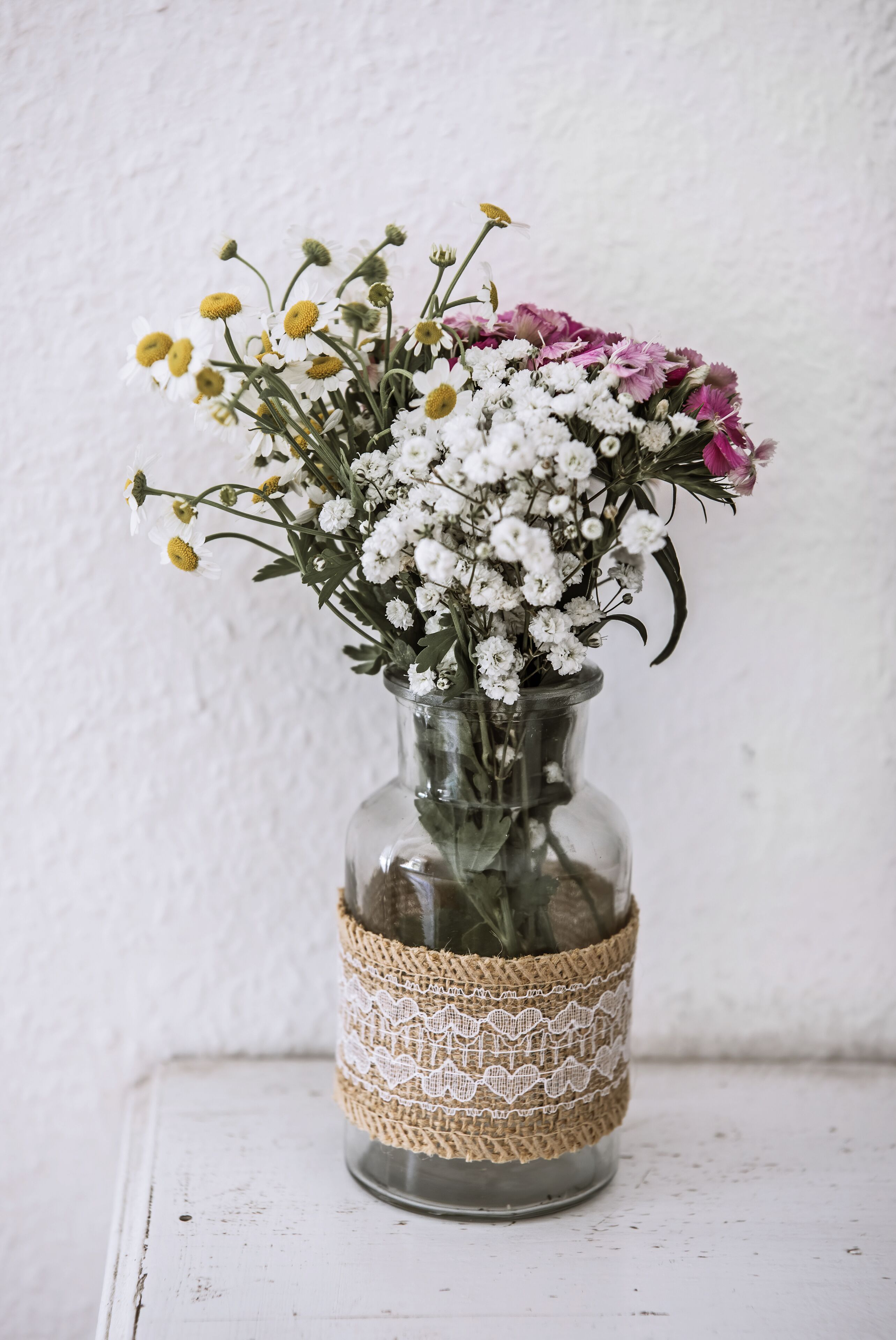 Un florero de vidrio transparente con una banda de arpillera y encaje, lleno de flores blancas y rosas, sobre una superficie blanca frente a una pared blanca.