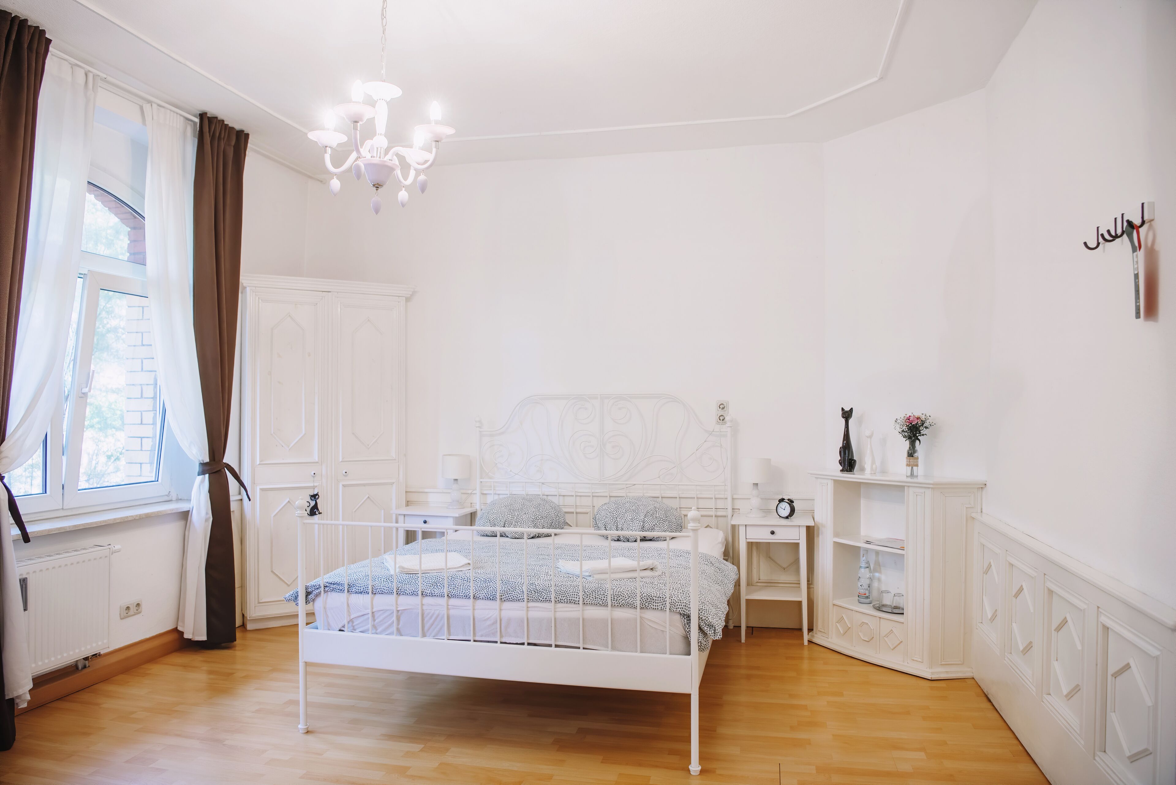 Une chambre à coucher avec un lit en métal blanc, une table de chevet, une commode, une coiffeuse et une fenêtre avec des rideaux.