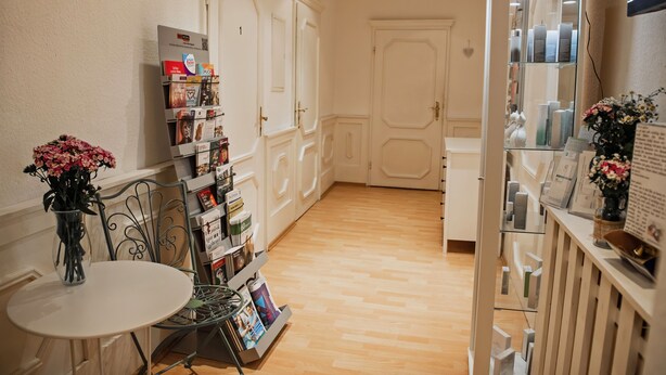 A hallway with a white door, a small table with a vase of flowers, and a shelf with various items. - Pension Bertha (Jena)