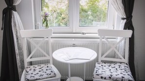 A room with two white chairs with patterned cushions, a small round table with papers, and a window with curtains. - Pension Bertha (Jena)