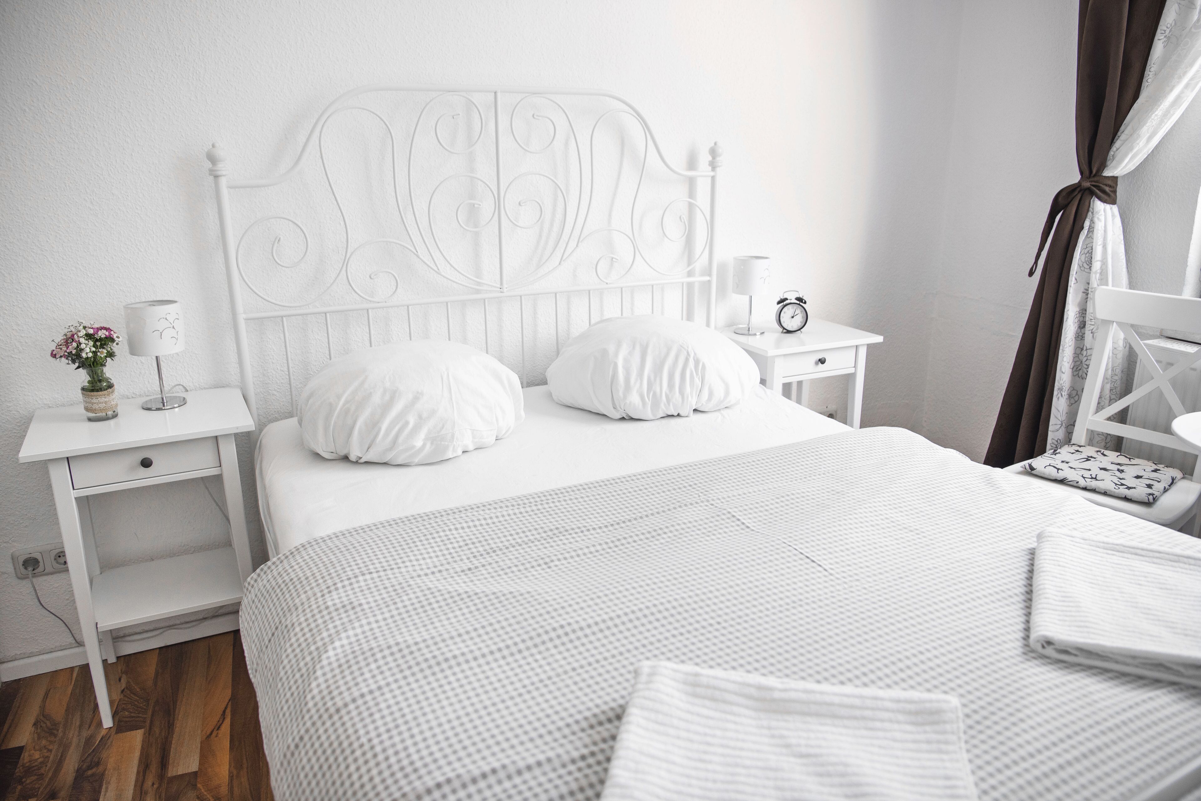 Una cama bien tendida con ropa blanca, cabecero de metal blanco, mesita de noche con lámpara y un pequeño reloj.