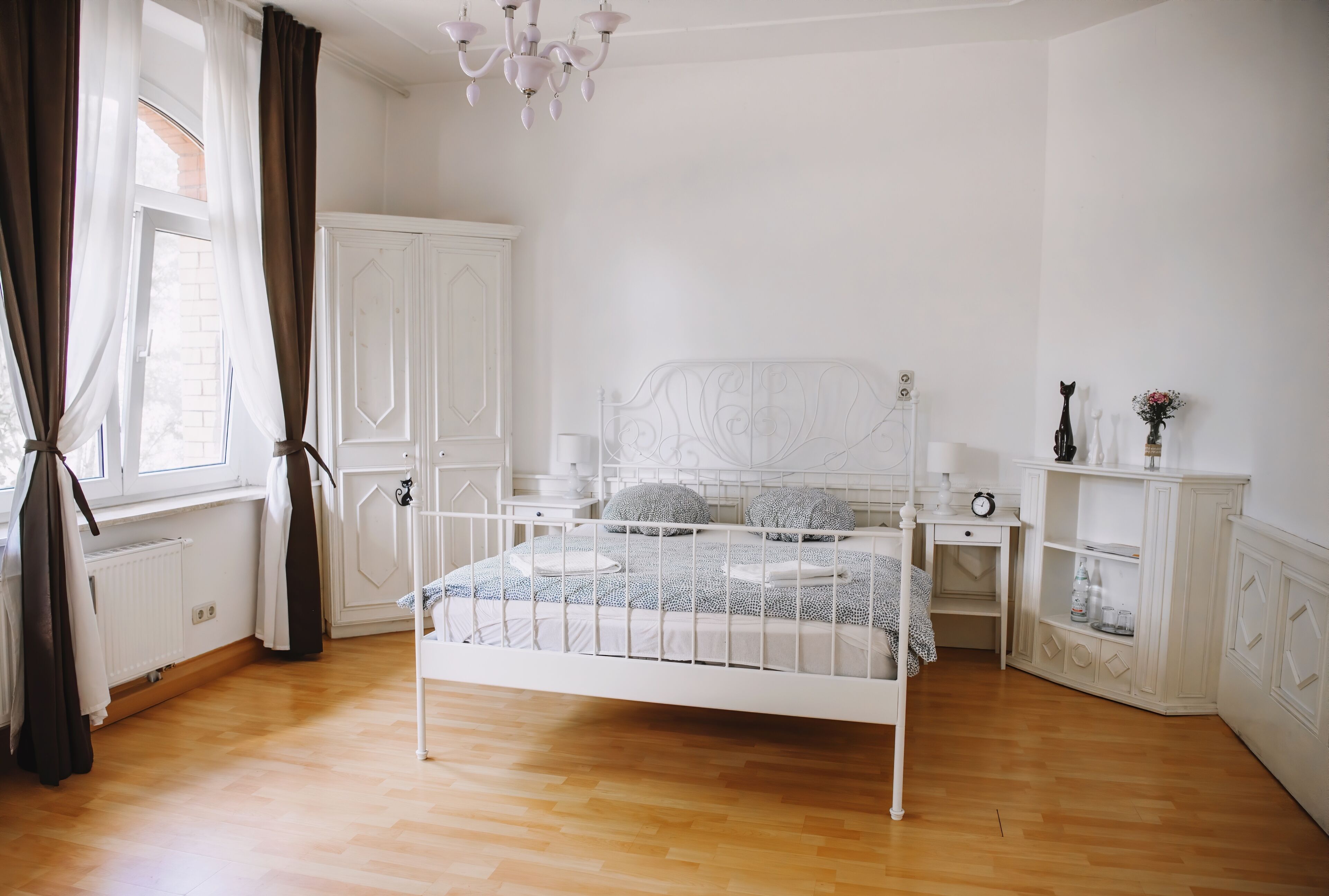 Un dormitorio blanco con estructura de cama de metal, suelo de madera, armario blanco, mesita de noche con lámpara y ventana con cortinas.