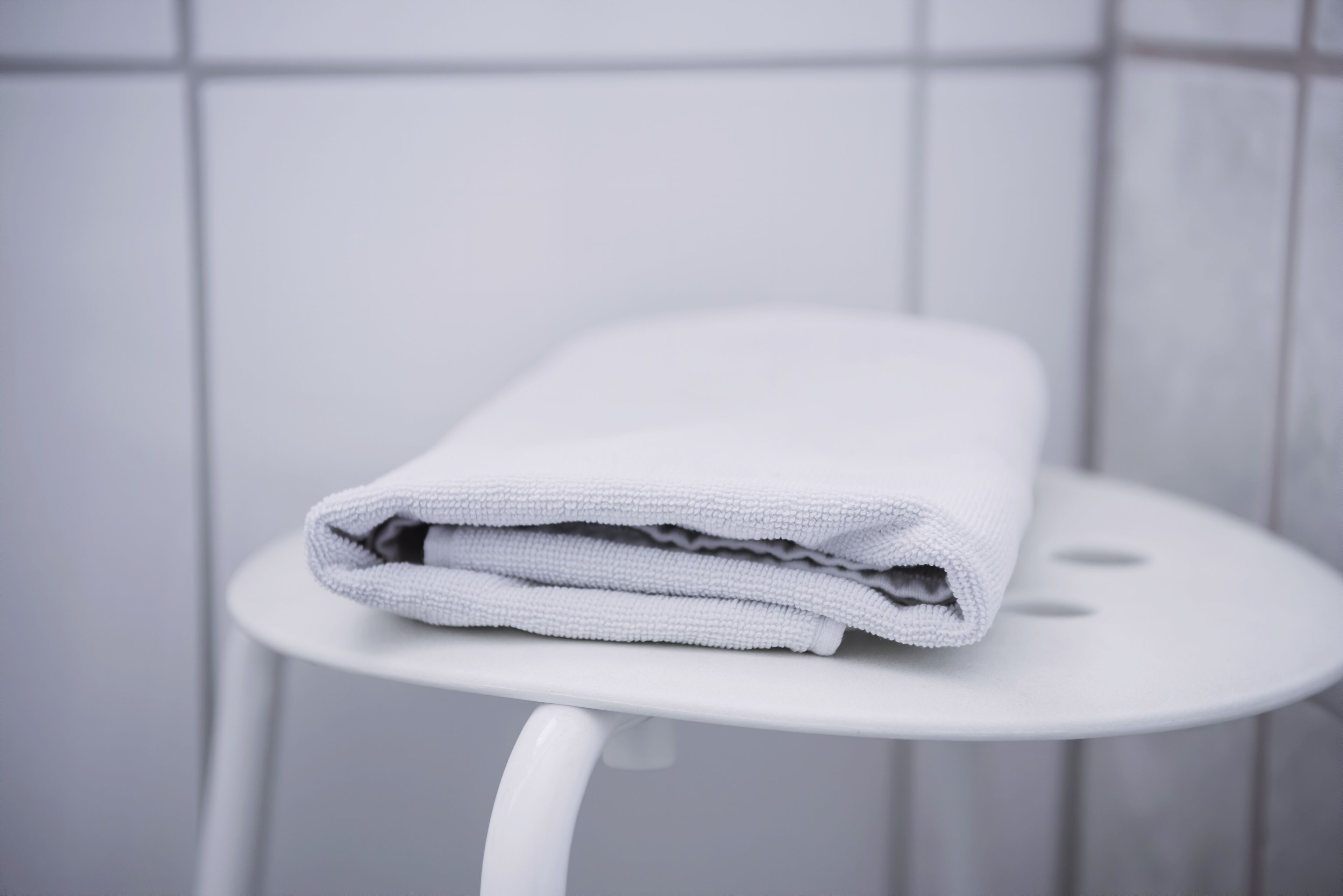 A white towel folded on a white stool in a bathroom with white tiles.