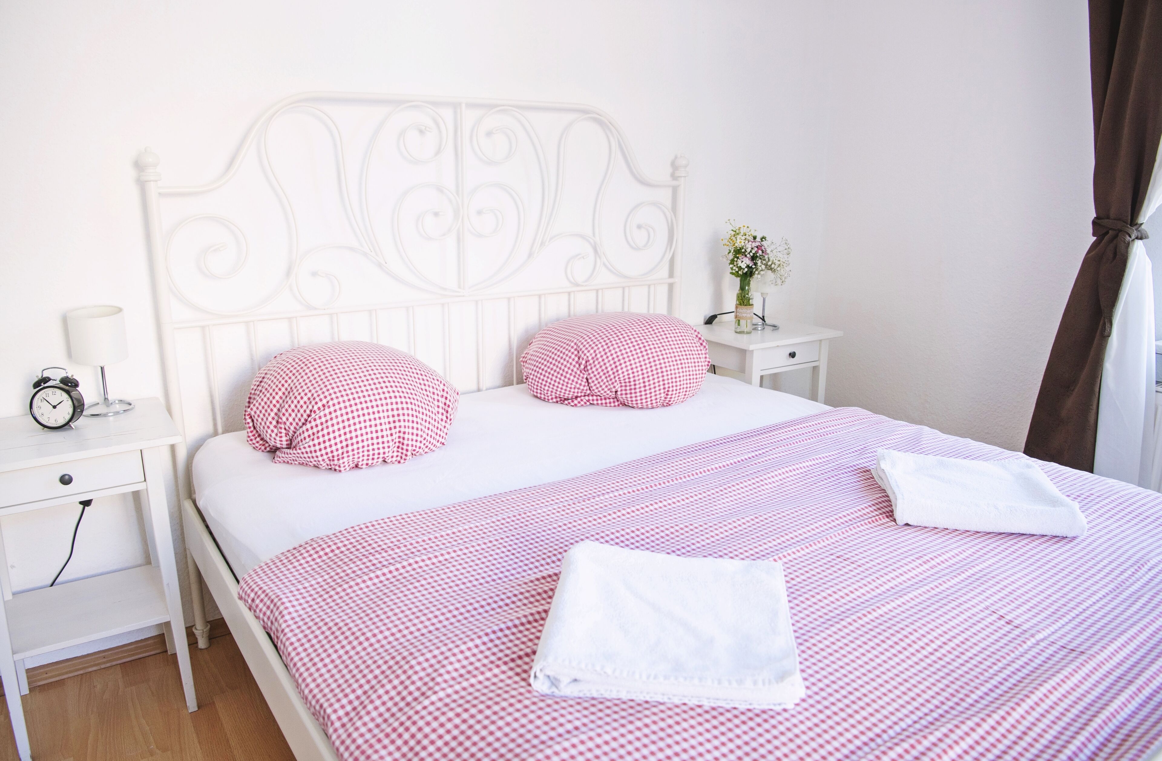 A neatly made bed with a checkered bedspread and pillows, a bedside table with a lamp and a clock, and a small nightstand with a vase of flowers.