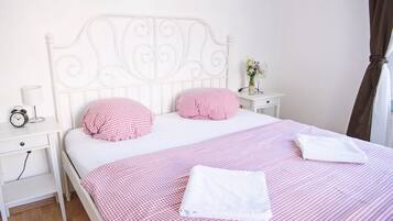 A neatly made bed with a checkered bedspread and pillows, a bedside table with a lamp and a clock, and a small nightstand with a vase of flowers.