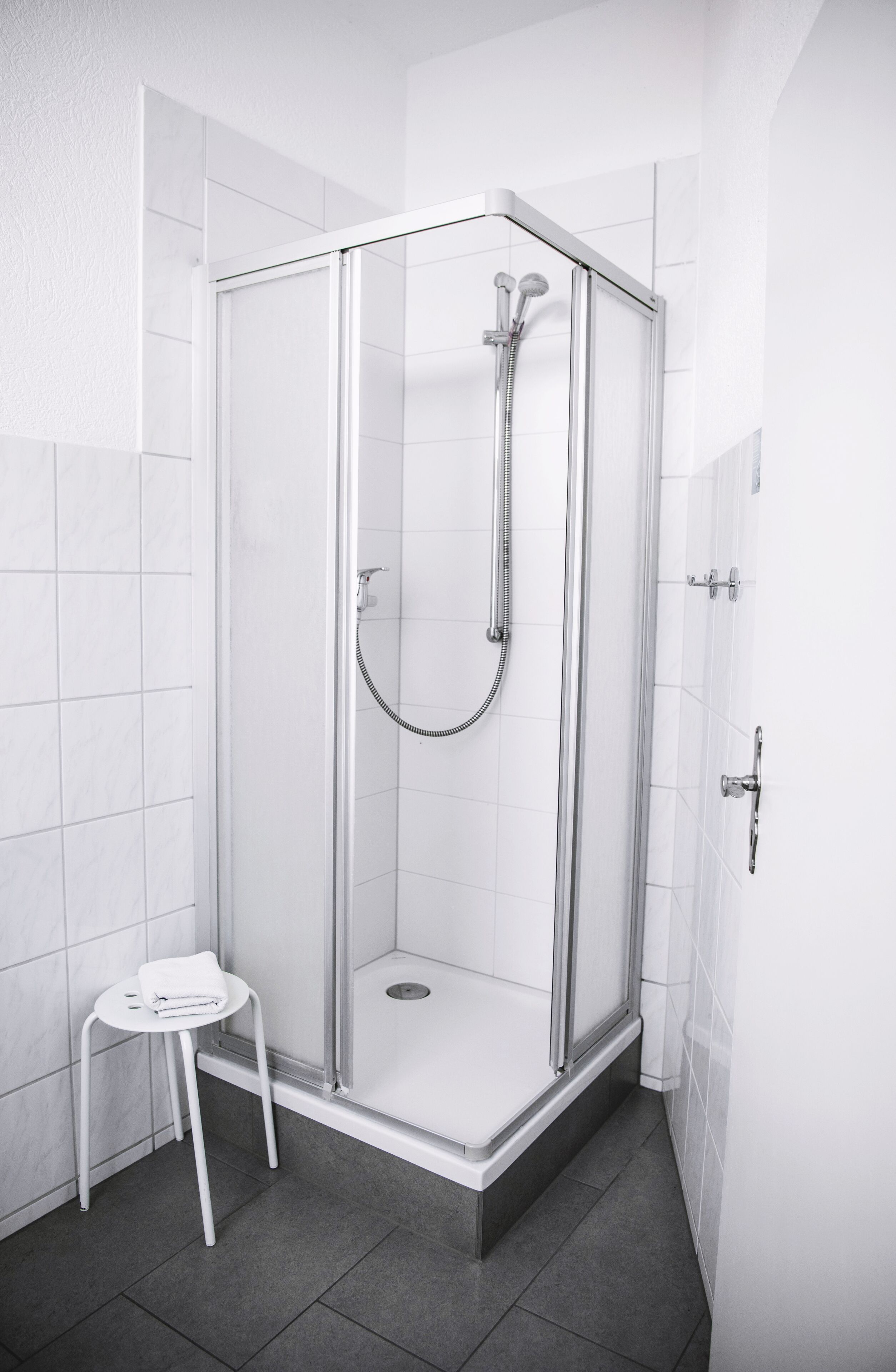 Box de chuveiro moderno com porta de vidro, chuveiro manual e um banquinho branco pequeno.