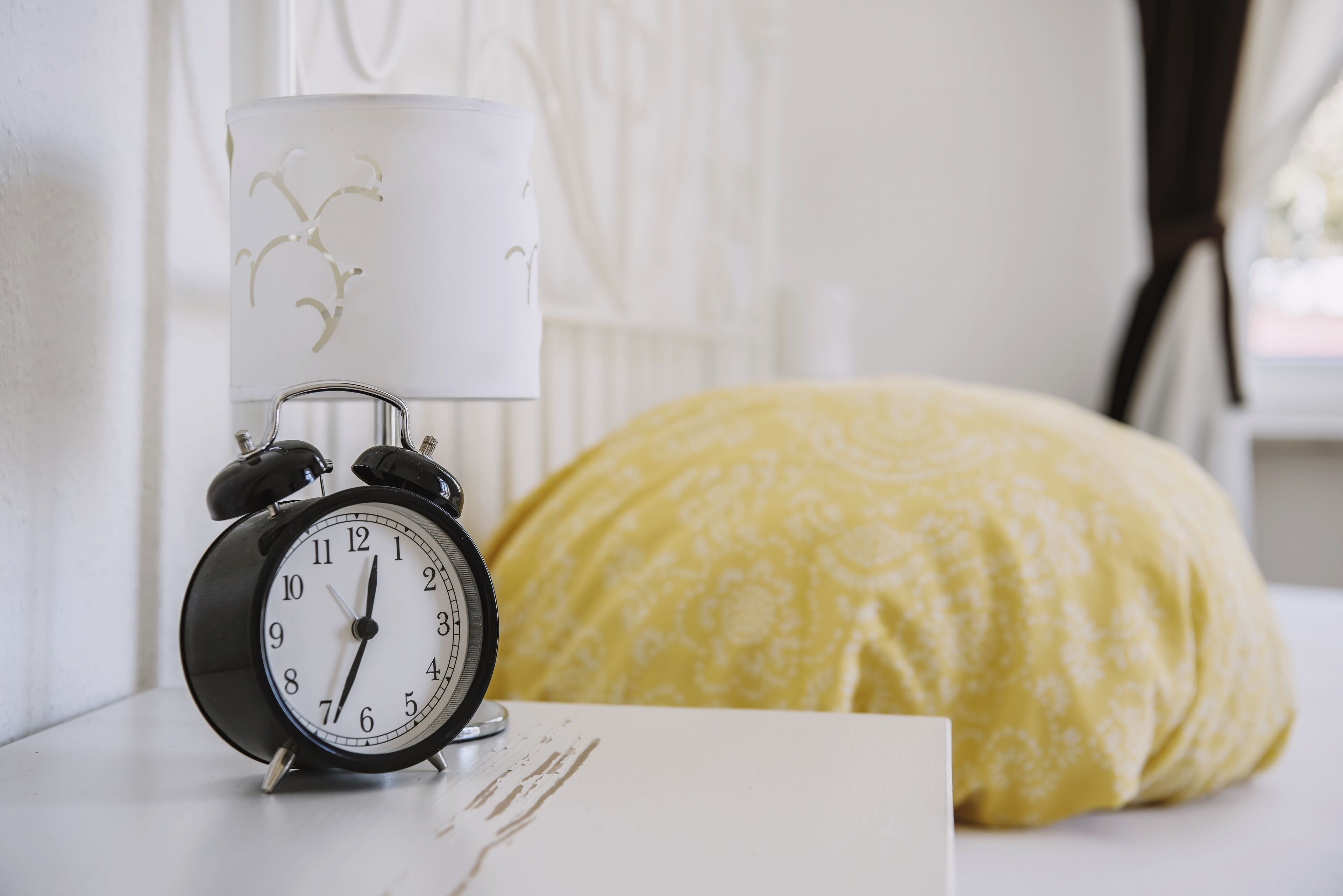 A white bedside table with a black alarm clock and a white lamp with a floral pattern.