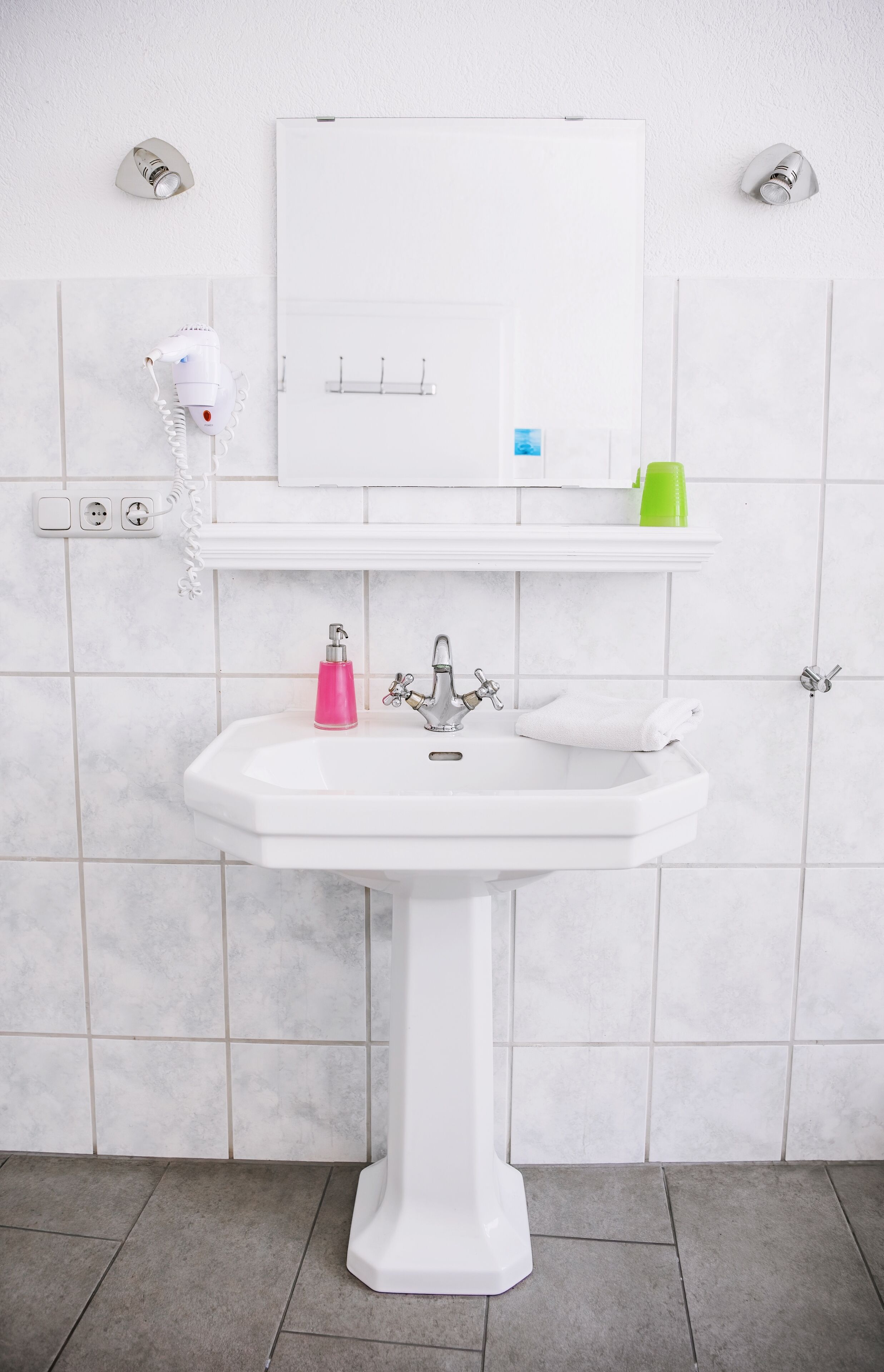 A white bathroom with a pedestal sink, a mirror above it, and a hairdryer on the wall.
