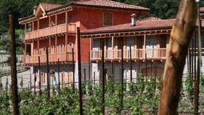 A two-story building with a red facade and a balcony, surrounded by greenery and a mountainous backdrop. - Maso Fiorini (Isera)