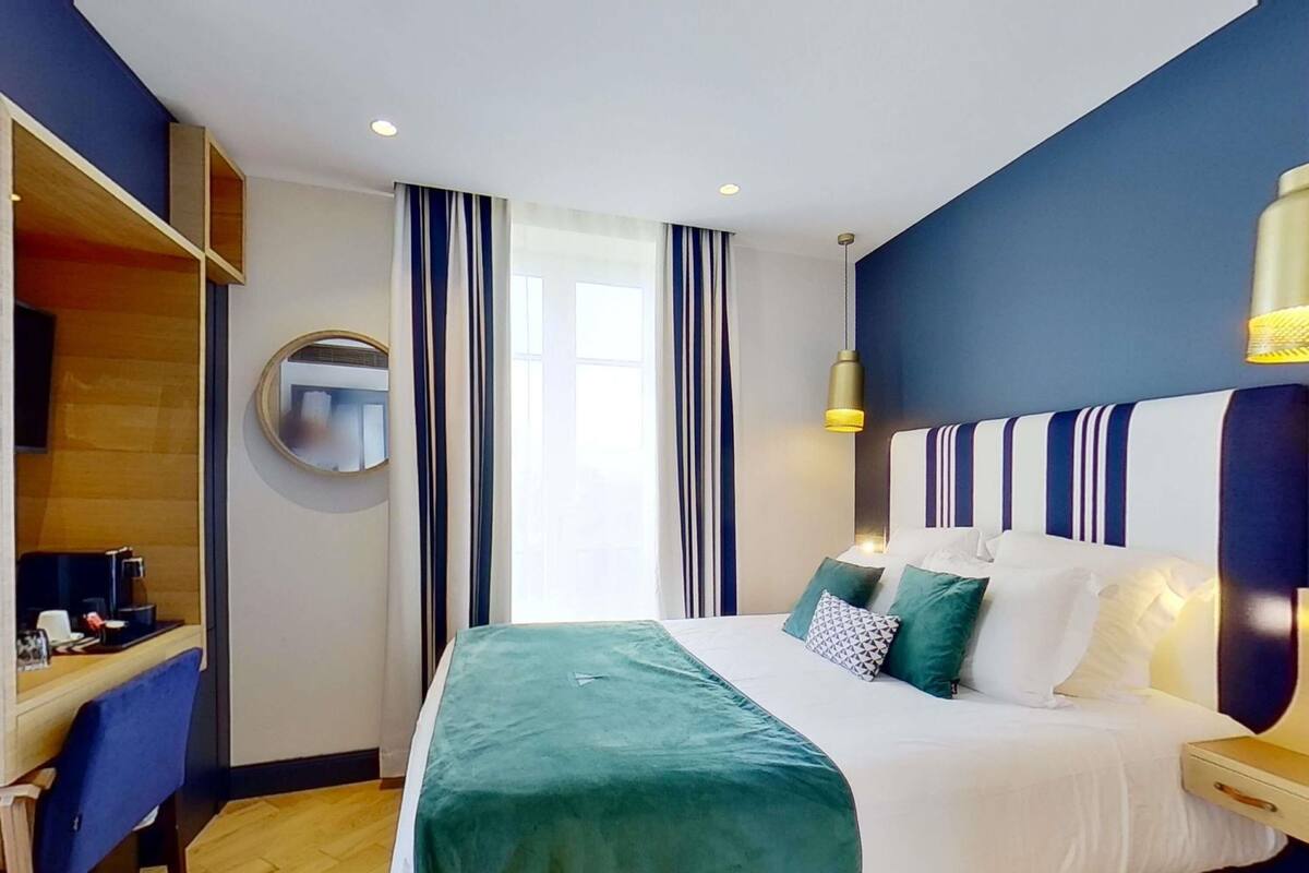 A hotel room with a bed, a desk, a mirror, and a window with curtains.