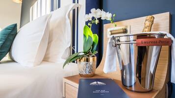 Una cama bien hecha con sábanas blancas, un florero decorativo con una planta, un cubo para champán y un folleto del servicio de minibar sobre una mesita de noche de madera.