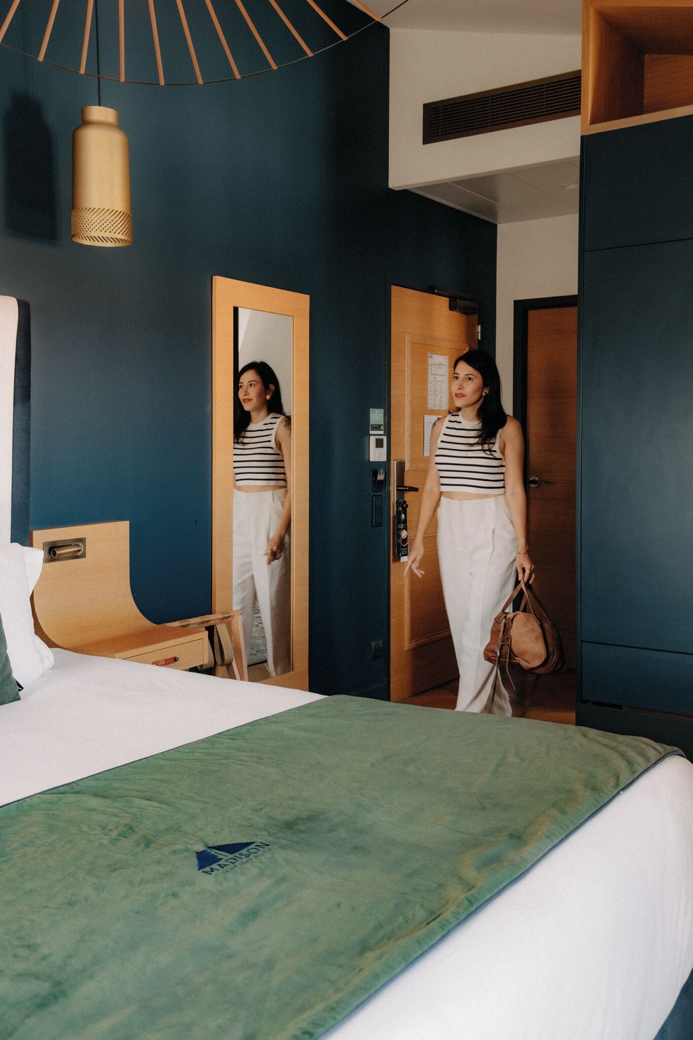 A hotel room with a bed, a wooden door, a mirror, and a hanging light fixture.