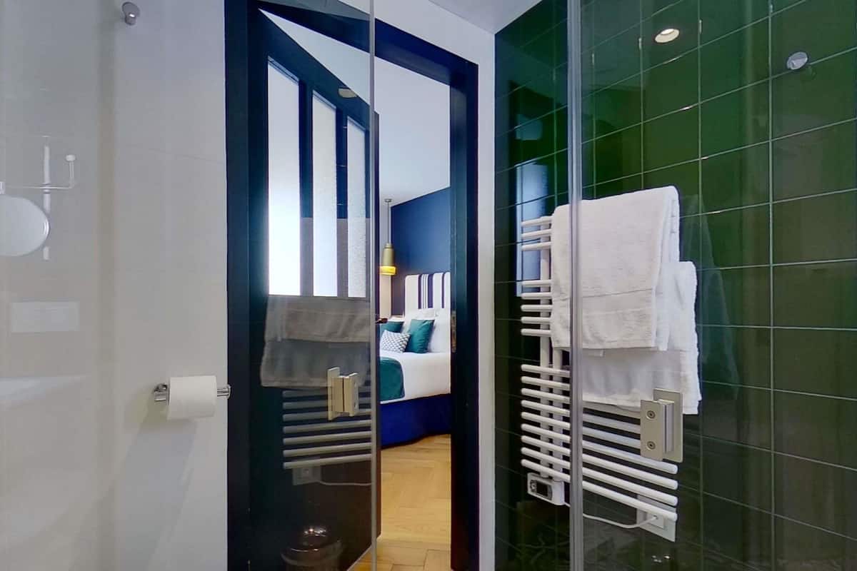 A modern bathroom with a glass shower enclosure, a toilet, and a visible bedroom with a bed and a nightstand.