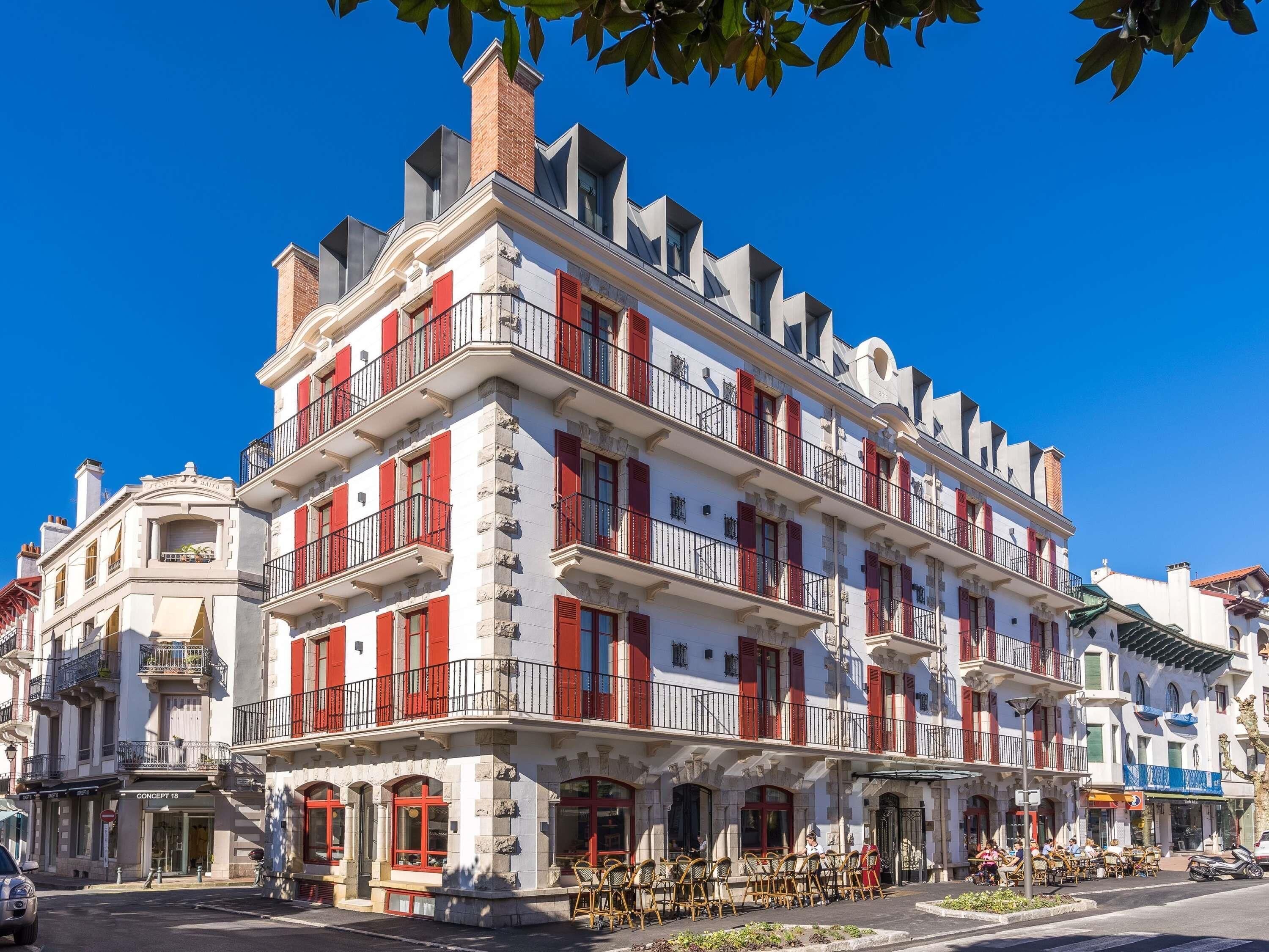 Un immeuble à plusieurs étages doté de balcons, d’un café situé au rez-de-chaussée et d’une devanture visible depuis la rue.