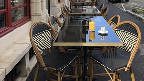 An outdoor café with a long table and wicker chairs, set against a building with classical architecture.