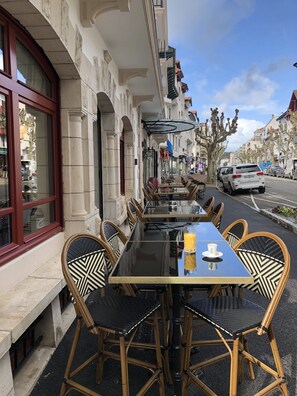 An outdoor café with a long table and wicker chairs, set against a building with classical architecture. - MADISON Saint Jean de Luz - Handwritten Collection (Saint-Jean-de-Luz)