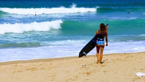 A beach with clear blue water and waves, a person holding a surfboard, and a sandy shore.