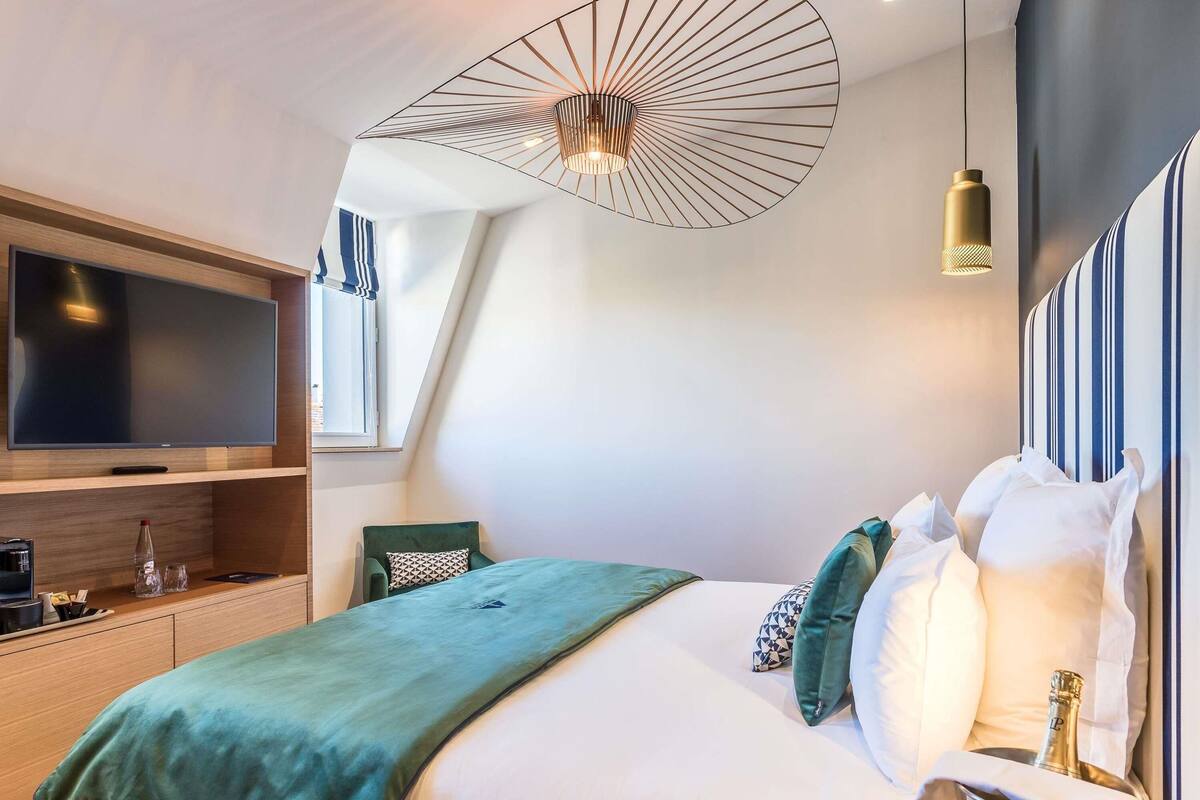 Ett modernt hotellrum med en stor platt-TV, en säng med många kuddar, en grön pläd och en unik taklampa.