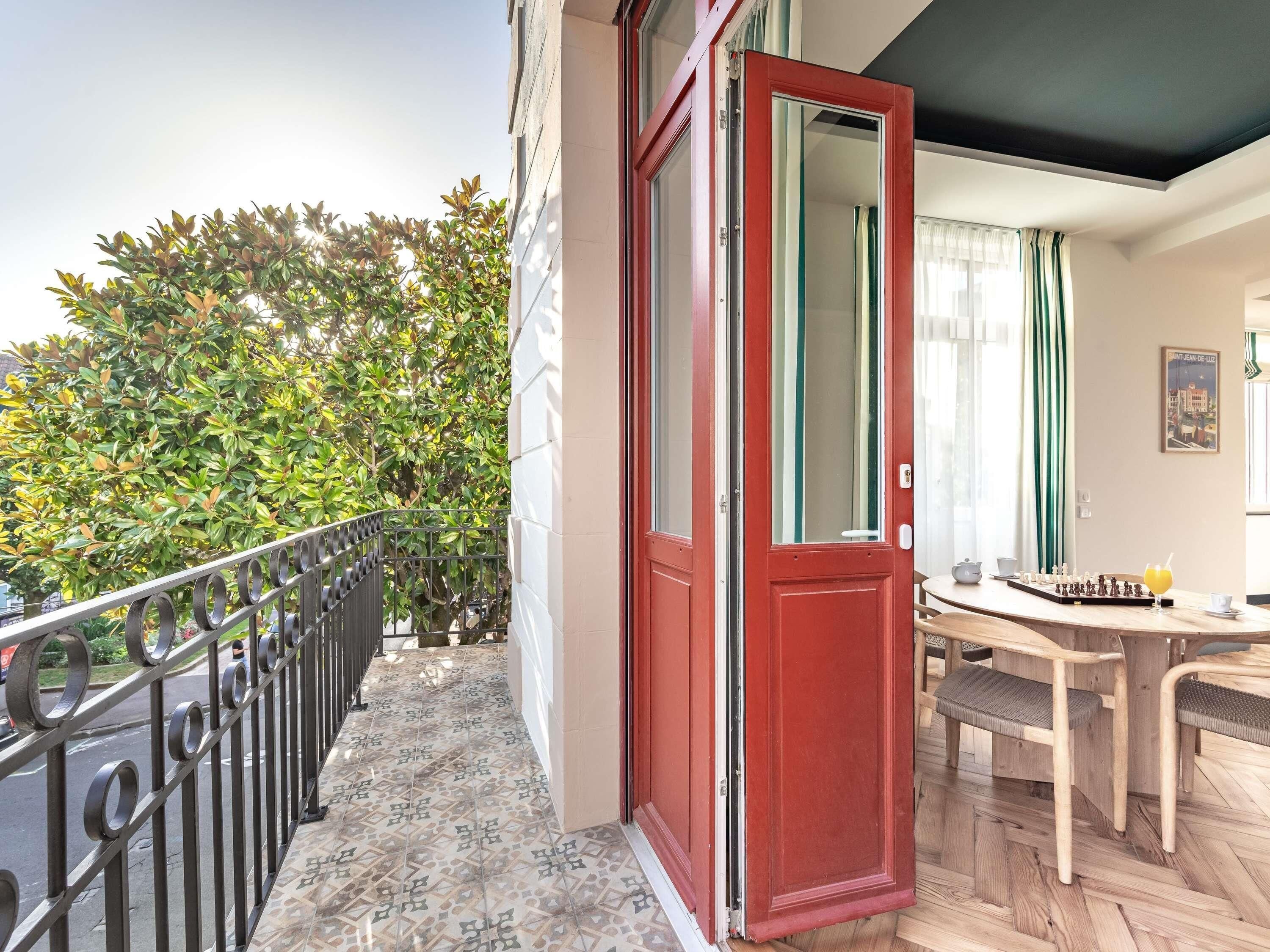 Un balcon avec une balustrade en métal, une porte rouge et une vue sur un arbre verdoyant.