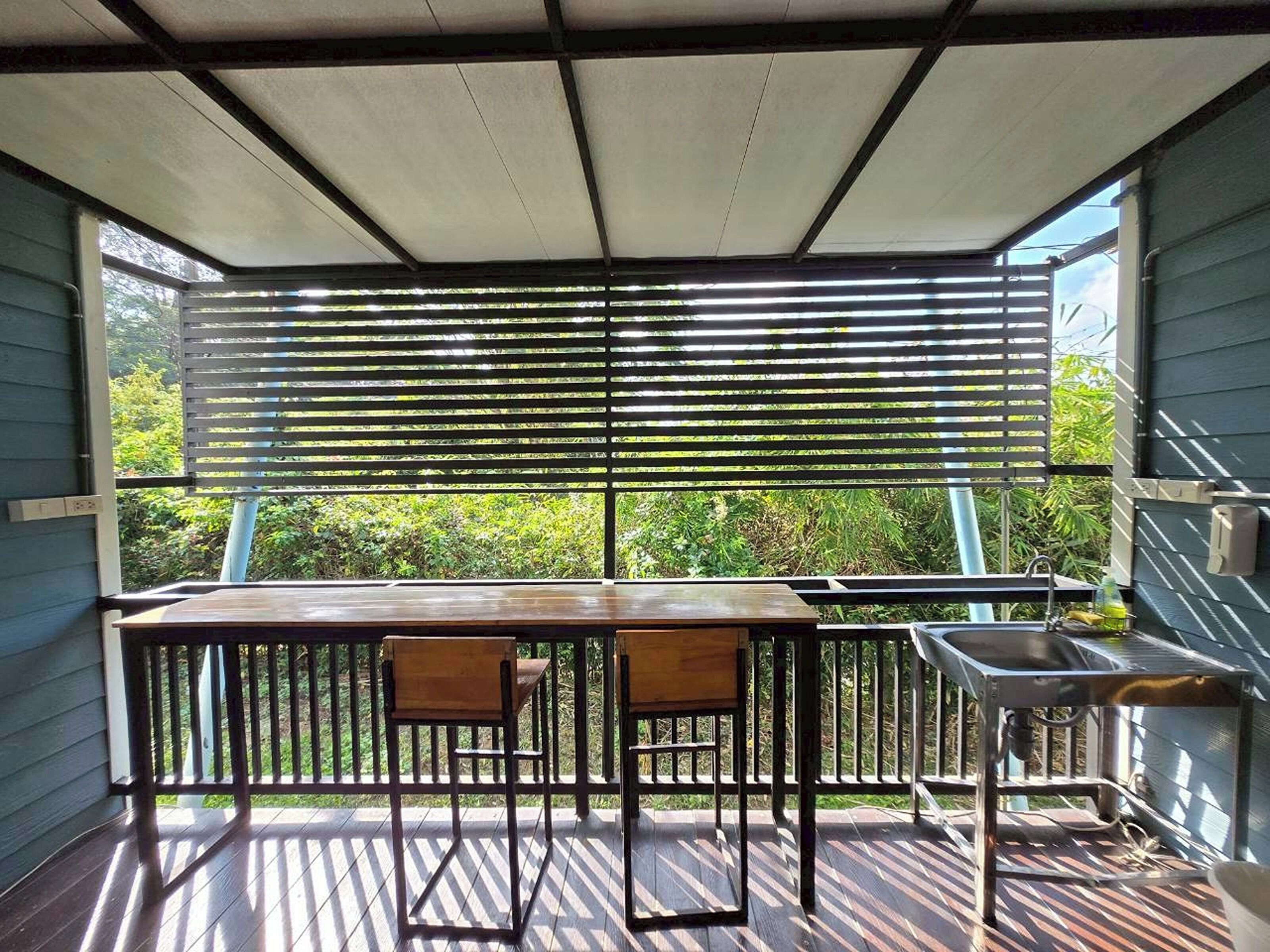 A covered outdoor area with a table and chairs, a sink, and a view of greenery.