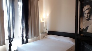Ein Hotelzimmer mit einem Bett, Vorhängen, einem Nachttisch mit Lampe und einem gerahmten Bild an der Wand.