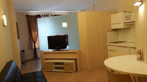 Ein gemütlicher, kleiner Wohnraum mit Fernseher auf Holzfuß, einem kleinen Esstisch und einer Küchenzeile mit Mikrowelle.